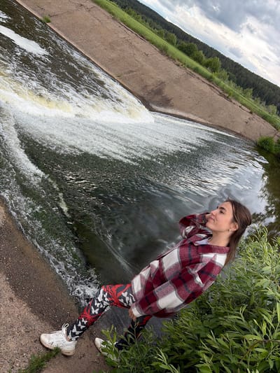 The image, taken in what appears to be Nizhnii Vishur, Russia, depicts a young woman standing near a small dam or waterfall. She is positioned on the bank, close to the edge where the water flows over. The woman is wearing patterned leggings, white sneakers, and a red and white flannel shirt. She has her hand near her face, looking down at the water. Lush green foliage surrounds her. Behind her and to the left is a concrete structure that directs water. Beyond the concrete structure, the landscape includes trees and a cloudy sky. The water flows over the edge of the concrete structure, creating a waterfall effect.