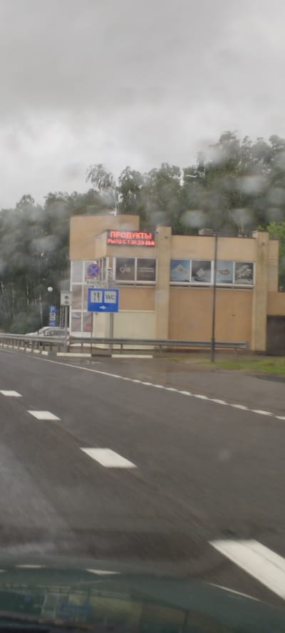 The image shows a roadside scene on a cloudy day, with rain streaks visible on the foreground, suggesting it was taken from inside a vehicle. In the background, a building with beige and brown facade stands beside a wooded area. A red LED sign on the building displays "ПРОДУКТЫ" (Products) and "РЫТО С 7.30 ДО 23.0" (Open from 7.30 to 23.0). Below this sign, a blue sign with white pictograms indicates "WC" with an arrow pointing to the right. Several windows on the building display images, including one with the "glo" logo. A metal barrier runs along the side of the asphalt road, which has white lane markings. A street lamp is also visible. The overall setting appears to be a commercial establishment along a highway in the vicinity of Верцялішкаўскі сельскі Савет, Belarus.