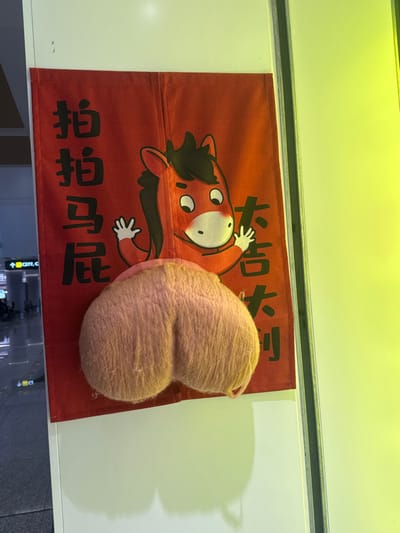 An indoor scene in Ürümqi Diwopu International Airport, China, displays a red fabric banner mounted on a white vertical surface. The banner features black Chinese characters and a cartoon horse. On the left, the characters "拍拍马屁" (pāi pāi mǎ pì, meaning "patting a horse's backside" or "to flatter") are vertically arranged. On the right, "大吉大利" (dà jí dà lì, meaning "great fortune, great prosperity") are also vertically arranged. A cartoon horse with a red body, black mane, and white muzzle is depicted with its arms raised. Directly below the horse's torso, a light-brown or peach-colored, furry, dual-lobed plush object, resembling a scrotum, is physically attached to the banner. The background on the left reveals a glass partition overlooking a large, brightly lit public area, with a distant sign displaying "C27L." A prominent vertical bright yellow-green light source illuminates the right side of the image.