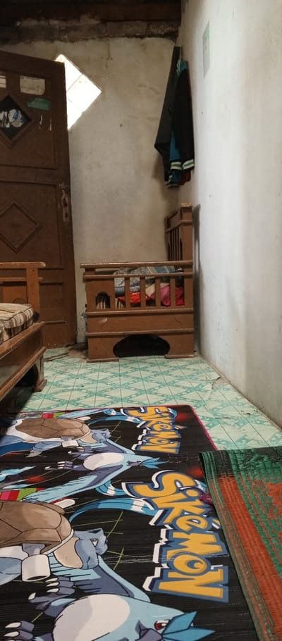 This image depicts the interior of a room in Weintiapa, Liberia. The floor is covered with light blue and white geometric patterned tiles, with visible signs of wear and some missing sections exposing a darker underlayer. Two mats are present on the floor: a large black mat featuring a "Pokemon" graphic, including characters such as Blastoise and Articuno, occupies the foreground and center, while a smaller woven mat with green and orange patterns is partially visible on the right.

The walls are painted an off-white or cream color, showing signs of aging, including stains and minor cracks. At the top of the corner where the back and right walls meet, some brickwork is exposed. A dark brown wooden door, featuring two diamond-shaped recessed panels, is positioned on the left side of the frame and is partially open, revealing a bright light source from the exterior. Various stickers or papers are affixed to the surface of this door.

On the far left, a portion of a wooden bed frame with a striped mattress (white and dark stripes) is visible. Against the back wall, positioned to the right of the open door, is a dark brown wooden crib or small bed with vertical slats and a carved base. This crib contains several folded textiles or clothes in various colors, including blue, pink, and red. Several garments, including a dark jacket and a blue shirt, are hanging from a hook on the right wall, near the corner. No people are present in the scene.