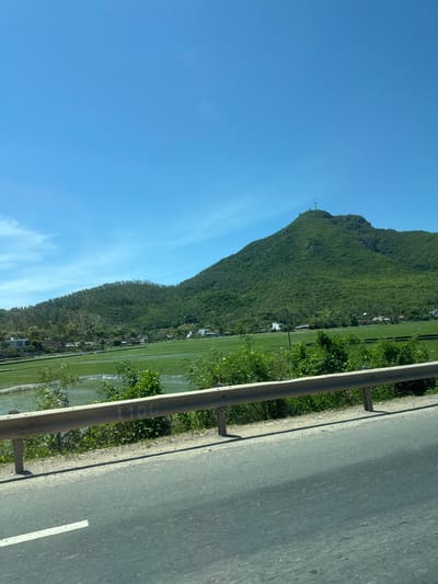 The image depicts a scenic view of a lush green mountain range under a clear blue sky. In the foreground, a grey asphalt road with a white lane marker runs diagonally across the frame. A metal guardrail, weathered and grey, separates the road from a vibrant green field that appears to be a rice paddy. Beyond the field, a cluster of houses with red and white roofs is visible, nestled at the base of the mountain. The mountain itself is densely covered in verdant vegetation, with some rocky outcrops visible. A telecommunications tower stands prominently on the summit of the highest peak. The scene suggests a rural or semi-rural location, likely in the vicinity of Tuy Hoa, Vietnam, known for its picturesque landscapes that often combine mountains, agriculture, and coastal elements. There are no people or animals visible in the frame.