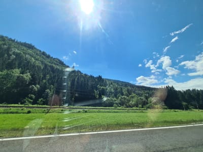 The image shows a sun-drenched landscape with a bright sun and blue sky, featuring a lush green mountain covered in dense coniferous trees on the left. A railway track runs horizontally across the middle ground, bordered by a vibrant green grassy field. The foreground consists of a paved road with a white line marking its edge. The scene is likely captured from a moving vehicle, indicated by the reflections on the lower part of the image and the perspective. The location is specified as Wildbad Einöd, Austria, suggesting a rural or mountainous area known for its natural beauty and possibly recreational activities. There are no people or animals visible in the frame. The overall impression is one of natural beauty and serenity on a clear, sunny day.