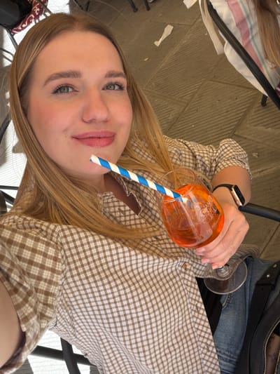 A young woman, possibly in her early to mid-20s, is taking a selfie while seated at a table. She is wearing a brown and white checkered shirt, jeans, and a black watch. She is holding a wine glass filled with an orange liquid and a blue and white striped straw. The background appears to be an outdoor seating area with stone flooring. The location may be San Gimignano, Italy, given the overall aesthetic.