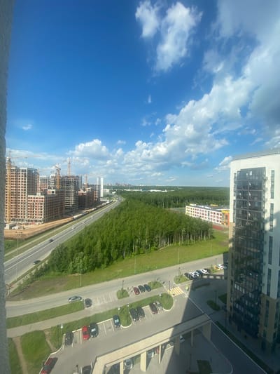 The image captures a wide aerial view of a developing urban area on a bright, sunny day in Kazan, Russia. The foreground is dominated by a large, multi-story residential building under construction on the left, with several construction cranes visible. Adjacent to the construction site is a multi-lane road with a few cars driving. Beyond the road, a dense green forest stretches across the landscape. In the middle ground, there is a newly constructed school building with a colorful facade, and further in the distance, more residential buildings and infrastructure can be seen, hinting at further development. The sky is a vibrant blue with scattered white clouds. The scene suggests a modern city expanding into a green, natural area.
