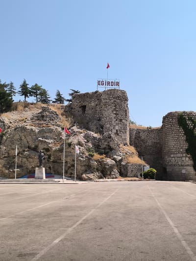 A statue of a man stands on a pedestal in front of a rocky hill. The hill has ruins of a castle on top, with a sign that says "EĞİRDİR." The castle is made of stone and some parts are covered in ivy. In front of the hill, there is a paved area with parking lot lines. Several flags are flying on poles. The sky is clear and blue. The environment appears to be an outdoor public space in the city of Eğirdir, Turkey.
