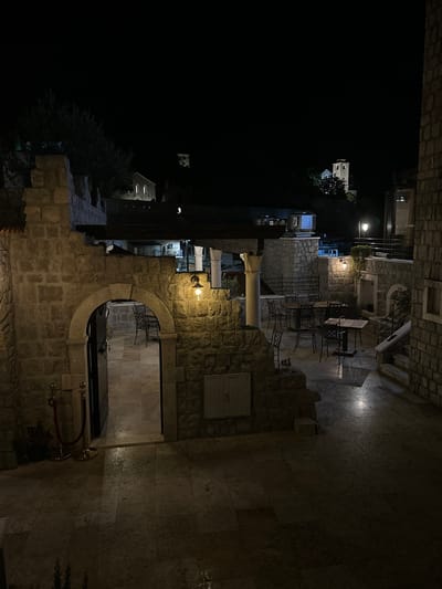 The image depicts an outdoor courtyard at night, illuminated by artificial lights. The architecture suggests a historic setting, with rough-hewn stone walls and an arched entryway. Several tables and chairs are arranged in the courtyard, indicating it is likely a dining or seating area for a restaurant or cafe. In the background, illuminated buildings, including what appears to be a church tower with a clock, are visible on a hillside against a dark sky. The ground is paved with large stone tiles, and some areas appear to be wet, reflecting the light. A velvet rope barrier with gold stanchions is positioned near the arched entrance. The overall scene is tranquil and atmospheric, characteristic of a European night scene. The location is identified as Bartula, Montenegro.