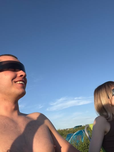 The image shows two people in an outdoor setting, likely a field or park, under a clear blue sky with some wispy clouds. The person on the left is a male, shirtless and wearing large black sunglasses that cover his eyes. He is smiling and looking to the right. His chest and shoulder are visible, with some light chest hair. The person on the right is a female, with blonde hair and also wearing sunglasses. She is partially visible from the shoulder up, with her head turned to the left. In the background, there are some tents and possibly a vehicle, suggesting a camping or outdoor event. The overall lighting suggests it is daytime, possibly late afternoon due to the warm tones on the skin. The location is identified as Povarovka, Russia.