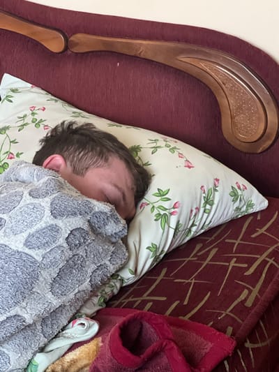 An overhead shot shows a person sleeping or resting comfortably indoors. The individual, whose face is covered by a blanket, is lying on their side, revealing the back of their head with short, dark hair and an ear. They rest on a white pillow adorned with a pattern of green leaves and delicate pink flowers.
A plush, textured blanket, in shades of grey and white with a relief pattern resembling large, rounded shapes, covers the person from the neck down. Surrounding the sleeper is rich, dark red (maroon) upholstered furniture, possibly a bed headboard or sofa back, featuring a subtle pattern of intersecting light gold lines. To the left, a decorative, polished wooden element with a carved or textured inset pattern is integrated into the furniture design, along with a smaller wooden piece below it. On the right side of the frame, alongside the patterned upholstery, are other textiles including a red, fuzzy blanket or fabric, and some crumpled white fabric with a similar floral pattern to the pillow.
The scene conveys a sense of peaceful rest. No specific time of day is evident, but the activity suggests sleep or a nap. There is no visible text. The setting is an indoor space, consistent with a home environment in Cherliany, Ukraine.