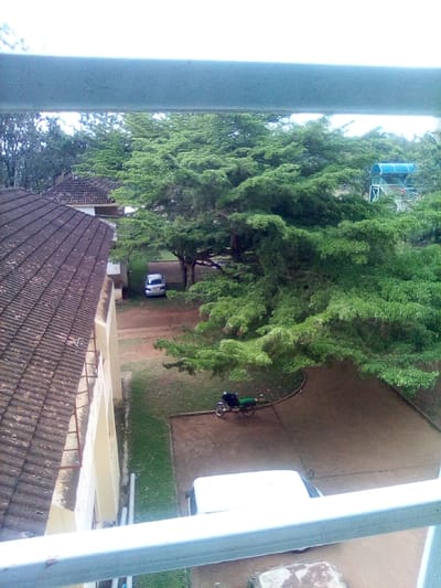 The image depicts an elevated view of an outdoor scene in West Sakwa ward, Kenya. A light-colored railing partially obstructs the top and bottom of the frame. In the left mid-ground, a building with a tiled, reddish-brown roof and light-colored walls is visible. A silver sedan is parked on a dirt path beside this building. Extensive green foliage from numerous trees dominates the central and right portions of the frame, with an overcast sky visible above. In the upper-right background, a multi-tiered structure with a blue canopy or roof is partially visible amidst the trees. A green and black motorcycle is parked on a dirt or paved path in the lower-mid ground, adjacent to a grassy patch. The white roof of another vehicle is partially visible in the bottom-right foreground, with a large rock or concrete block to its immediate right. No individuals are present, and no active interactions or specific actions are discernible.