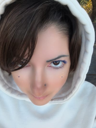 A medium close-up, high-angle portrait features a person with fair skin, dark brown hair, and brown eyes, looking directly upwards into the camera. The individual wears an off-white or cream-colored hooded sweatshirt, with the hood pulled up and framing the face. Dark brown hair falls forward, partially obscuring the left side of the forehead and right eye (viewer's left).

The person's eyes are prominent, with bright blue mascara applied to the lower lashes of both eyes and blue eyeliner visible above the upper lashes of the right eye (viewer's right). Small red veins are visible in the whites of both eyes, particularly in the inner corners. The eyebrows are dark and well-defined. A small, metallic silver stud piercing is present on the left nostril. Below the left eye, there are several small, irregularly shaped purple glitter flecks. Below the right eye, there are several small, gold-colored glitter flecks. The mouth is mostly obscured, with only the upper lip and a hint of the lower lip visible in a light pink hue.

The background is out of focus. To the viewer's upper right, indistinct yellowish-green foliage and dark shapes, possibly tree trunks or wooden structures, suggest an outdoor or partially outdoor setting. The lighting appears natural, illuminating the face from the front.