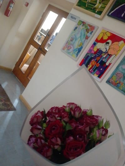 The image depicts an indoor scene, likely a hallway or common area, with several pieces of art displayed on a white wall. In the foreground, a bouquet of deep red roses is presented in a white wrapping. To the left, a wooden door with glass panels is visible, revealing a glimpse of an interior space with furniture. Further to the left, a red fire extinguisher is mounted on the wall, with a smaller red alarm device above it. The floor appears to be a light gray tiled surface, with a patterned rug in the lower left corner. The lighting suggests it is daytime, with light entering from the direction of the door. The overall impression is of a clean and decorated interior space, possibly in a public building or residence. The specific location in Nor Hachin, Armenia, is not evident from the visual information provided in the image itself.