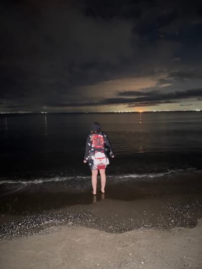 A person stands on a beach at night, facing the ocean. The person is wearing a bucket hat and a long-sleeved shirt with a red and white Japanese-style print of a cat. They are also wearing shorts with a red and white pattern. The ocean is dark and calm, with gentle waves lapping at the shore. In the distance, lights from a city are visible on the horizon. The sky is cloudy and dark, with some faint moonlight. The beach is covered in sand and small pebbles. The overall scene suggests a quiet and contemplative moment by the sea. It is highly likely this location is the coast of Chiba City, Japan, given the appearance of urban lights on the horizon across the water, characteristic of coastal cities in Japan.