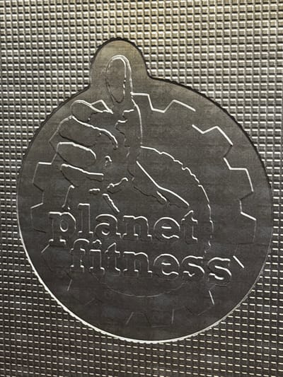 The image shows a close-up, overhead view of a metallic circular object with the text "planet fitness" embossed on it. The object is centered on a textured, grid-like surface. The object itself appears to be a charm or a pendant, with a ridged edge and a detailed embossed design. This design includes a silhouette of a person with their arm raised, forming a thumbs-up gesture, superimposed over a gear shape. The lighting highlights the embossed details, creating shadows that add depth. The background is a repeating pattern of small, raised squares, giving a tactile impression. There is no indication of the city of New Albany, United States of America within the image.