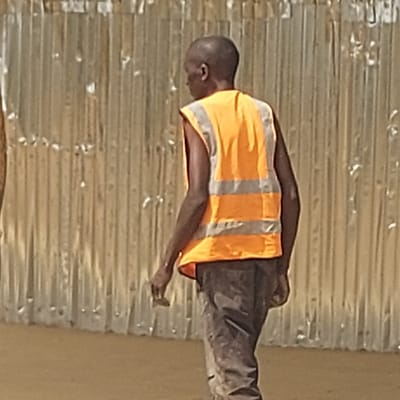 The image displays a person, likely an adult male, wearing a high-visibility vest in an outdoor setting. The original image appears to be rotated 90 degrees counter-clockwise, with the person standing upright on what would be the ground if corrected.
The individual has dark skin and a bald or closely shaved head. He is dressed in an orange high-visibility vest featuring two horizontal grey reflective stripes across the torso and one vertical stripe on each side. Underneath the vest, parts of a dark-colored garment are visible on his arms and neck. He is also wearing dark trousers, which appear somewhat soiled or dusty, suggesting manual work.
The person is captured from a slightly elevated perspective, looking downwards and to his right (the viewer's left if the image were reoriented). His posture suggests attentiveness or focus on something below him. His right arm is bent at the elbow, and his right hand is partially visible, fingers slightly curled. His left arm is mostly obscured by his body.
The background consists primarily of a textured surface that resembles corrugated metal sheeting, likely a wall or fence, running horizontally across the frame. This surface has a light brown or sandy color with various reflections and possibly some weathering. To the far right of the frame, there is a section of a lighter, plain-colored wall or surface.
The lighting indicates daytime conditions, with relatively strong illumination creating subtle highlights on the corrugated background and the reflective strips of the vest. No specific activity beyond standing and looking downwards is clearly depicted, but the attire suggests a work-related context, possibly a construction site, industrial area, or similar outdoor work environment. No text is visible in the image.