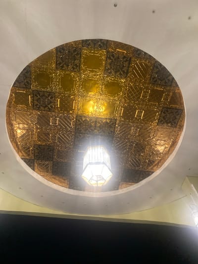 The image shows the ceiling of a building in Buea, Cameroon. The ceiling is made of a large, circular, recessed panel surrounded by a white perimeter. The panel is covered in gold, embossed tin tiles. The tiles form a pattern of squares, rectangles, and diamond shapes, with intricate, floral designs. A single, octagonal light fixture, with frosted glass panels and a metallic frame, hangs from the center of the panel. The fixture is lit, casting light on the surrounding tiles. The image is taken from a low angle, looking directly up at the ceiling. There are no people or objects in the image, other than the ceiling and the light fixture.