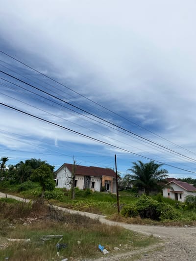 A low-angle shot depicts a cloudy sky over a rural landscape in Seunebobaru, Indonesia. Multiple dark power lines stretch across the upper portion of the frame, dividing the sky and obscuring some of the distant view. In the middle ground, a cluster of houses, some appearing under construction or in disrepair, are nestled amongst lush green vegetation and palm trees. A dirt road winds through the scene, with dry grass and scattered debris in the foreground. The overall impression is one of a developing or somewhat neglected rural settlement.