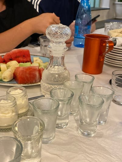 The image depicts a dining table in what is stated to be a establishment in Απολλωνία, Greece. The table is covered with a white tablecloth. In the mid-foreground, a clear, cut-glass decanter with a decorative stopper contains a transparent liquid. Arranged around the decanter are six small, clear, shot-style glasses, some exhibiting condensation. To the left of the decanter, a light blue plate holds multiple cut pieces of red watermelon and yellow banana slices. Further left, two small glass jars contain a white, creamy substance. To the right of the decanter, an orange, metallic, handled mug stands next to a stack of white plates. A blue plastic water bottle, with "Zaros" visible on its label, is positioned in the background. Two individuals are partially visible: on the left, a person's forearm, hand, and a portion of a black shirt are seen, with the hand extended towards the table; on the right, an arm in a dark blue shirt with white stripes on the sleeve is holding a black mobile phone. The background is out of focus, indicating an indoor setting.