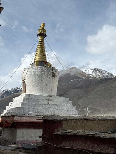 The image captures a white stupa with a gold spire in Tashi Dzom, China. A mountainous landscape featuring snow-capped peaks forms the backdrop. The sky above is partly cloudy. The stupa is partially connected to a building with a tiered roof, showing signs of age and deterioration. A few figures can be seen near the top of the stupa. Cables extend from the stupa's spire. The overall composition suggests a high-altitude, mountainous environment.