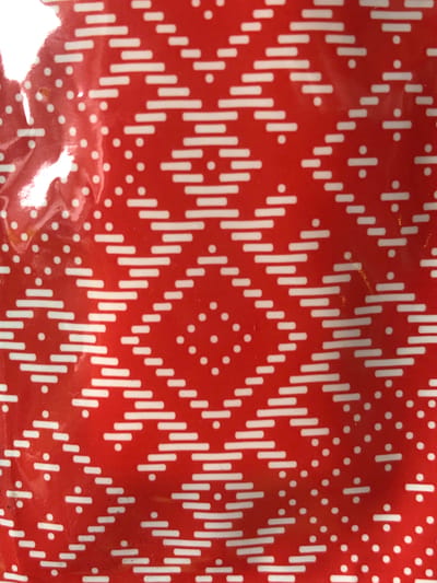 The image is a close-up, top-down view of a red surface with a repeating white pattern. The pattern consists of diagonal rows of short, horizontal white lines, creating a zigzag or diamond-like effect. Interspersed between these diagonal rows are scattered white dots, some appearing in clusters and others individually. The red surface has a glossy, reflective quality, with some areas showing glare and distortion, suggesting it might be a textured plastic or painted metal. The overall impression is of a decorative or patterned material, possibly from an object or packaging. There are no elements in the image that indicate a specific location in the city of Nachiki, Russia, or any people, actions, or interactions.