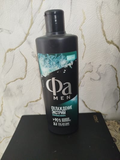 A black bottle of "Fa MEN" shower gel with "ARCTIC FRESHNESS" and "450 ml" text is positioned on a dark surface, possibly a book, with the year "2023" visible on it. The background is a textured wallpaper with a light color and gold-colored patterns. The scene is likely indoors, given the wallpaper. There is no information available to determine the location in the city of Буховіцкі сельскі Савет, Belarus.