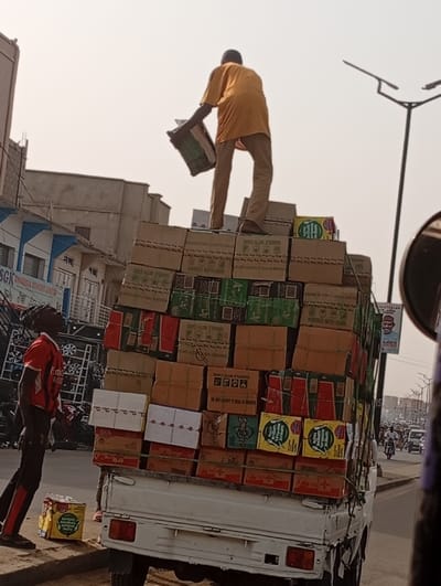 A truck overloaded with cardboard boxes, bearing labels like "HANDLE WITH CARE" and "SUPREME NOODLES PASTE." A man wearing a yellow shirt and light brown pants stands atop the pile, holding another box, while another man in a red shirt looks up at him from the ground. The scene is a bustling street with buildings, a street light, and other vehicles visible in the background. A poster of a man is attached to the back of the stack of boxes.