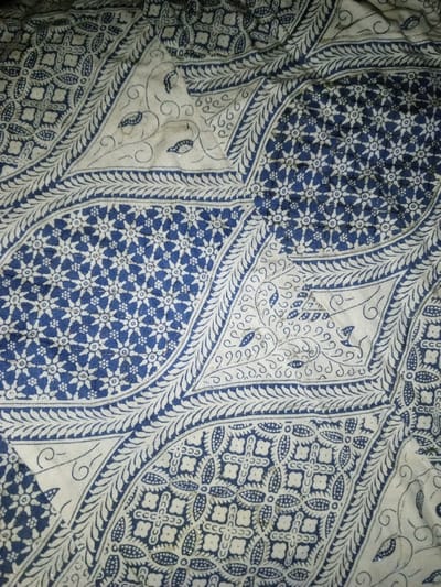This image displays a blue and white patterned textile, located in Na Sang, Laos. The fabric exhibits a complex design featuring distinct sections. Some areas showcase a dense repetition of small, white, six-petaled floral or star-like motifs against a dark blue background. Other sections present curvilinear patterns of blue foliage, leaves, or vine-like elements on a white ground, often bordered by intricate blue decorative bands with serrated or scalloped edges. A third type of pattern observed includes a geometric grid-like design with stylized blue shapes resembling interconnected squares or four-leaf clovers, set against white. Small, reflective, possibly silvery particles or glitter are visible scattered across various patterned areas, particularly within the blue sections. The textile appears to be a single piece of fabric, exhibiting some creasing and texture. No individuals, additional objects, or discernible actions are present within the frame, which is filled entirely by the textile.