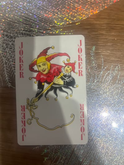 A single playing card, identified as a "JOKER" by the red lettering on its face, is positioned on a wooden surface. The card features an illustration of a jester character in a red and yellow costume, holding a golden staff with a ribbon. The card is oriented vertically, with the word "JOKER" printed in red letters along both the left and right edges. The wooden surface appears to be a table, and a silver, textured material, possibly a placemat or runner with a holographic sheen, is visible in the background and partially under the card. There is no indication of the city of Rosenovo, Bulgaria, or any specific environmental context beyond the table and placemat.
