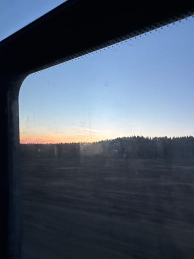 The image shows a view of the sky and landscape through a car sunroof. The sky transitions from blue to orange-yellow near the horizon, indicating sunrise or sunset. A dark line of trees is visible on the right side of the image, bordering the horizon. The dark frame of the sunroof is prominent in the foreground. The location, Krasnye Gorki, Russia, cannot be validated from the image content.