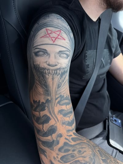 The image shows a close-up of a person's arm with a detailed tattoo. The tattoo depicts a scary, demonic-looking nun with a wide, toothy grin, blank white eyes, and a red pentagram on her forehead. The tattoo extends down the arm, featuring abstract, skeletal-like patterns. The person is wearing a black t-shirt with a subtle embossed logo. A dark seatbelt is visible across the chest, and a smartphone in a dark case rests on a surface to the right. The background appears to be the interior of a vehicle, with dark leather seating. The image was taken in Ivanski, Bulgaria.