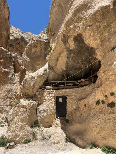 A low-angle shot shows the rock-cut St. Sergius and Bacchus church in Ma'loula, Syria. A dark metal door is visible, likely leading into the church interior, built into the rock. Wood support beams are adjacent to the entrance. The rock formation overhead creates a natural cave-like structure. The sky is visible through an opening in the rock. Vegetation grows on the rocky surface.
