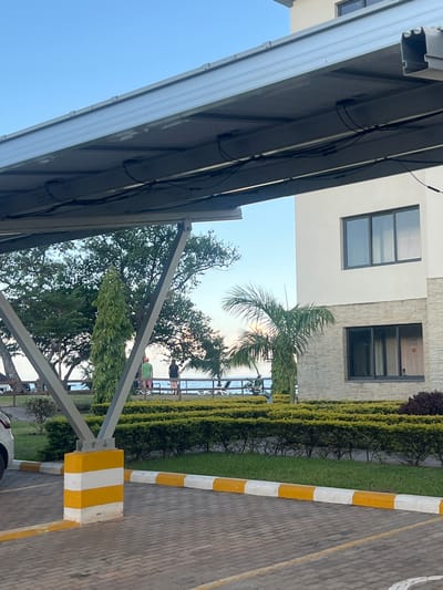 The image captures a view in Dowh, Malawi, likely from under a parking structure. The structure has a gray metallic frame and a reflective panel roof with attached cables. Adjacent to the structure is a building with two visible windows: one with three panes and the other, which has a textured facade around it, with two panes. A green hedge separates the building from a paved area marked with a yellow and white striped bollard and edge. In the background, trees and a body of water are visible, with figures of people discernible near the water.