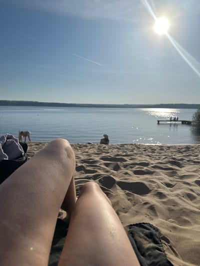 The image depicts a first-person perspective of someone sitting on a sandy beach in Matiushino, Russia. The view includes their legs, with one knee prominently visible. To the left is a body of water, possibly a lake or river, reflecting the sunlight. In the distance, there appears to be a pier or dock with figures on it. Further down the shoreline, two more individuals are visible. The sky is a clear blue with the sun shining brightly. A contrail from an aircraft stretches across the sky. The sand is textured, and there is clothing near the person's legs.