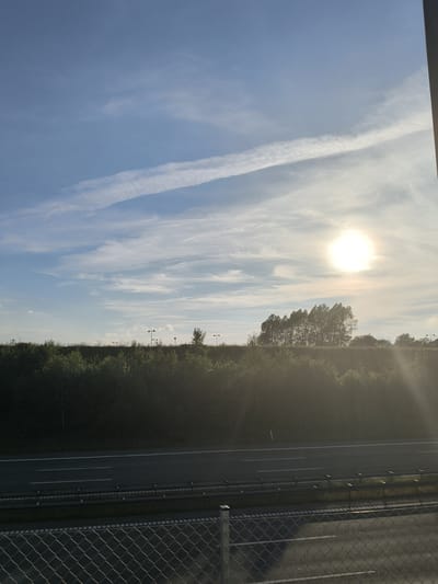 The image depicts a highway scene in Smidstrup, Denmark, under a partially cloudy sky. The sun is visible and bright, casting a glare. A dense line of trees and bushes forms a barrier between the highway and the sky. Several lampposts are intermittently spaced along the embankment behind the trees. In the foreground, a chain-link fence runs horizontally across the bottom of the frame, with a section of asphalt road beyond it. The highway itself is visible, with white lane markings. No people or vehicles are clearly discernible in the image.