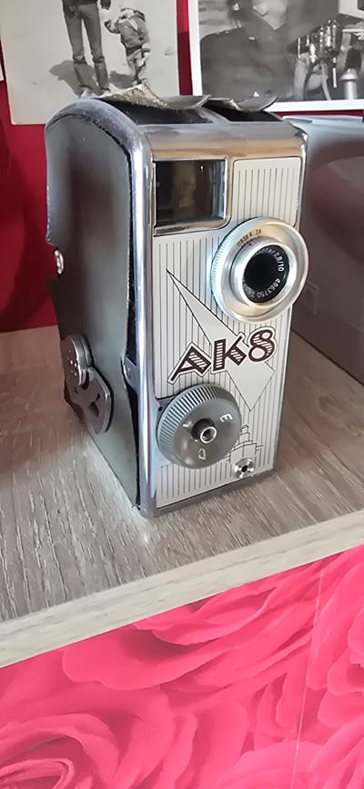 A vintage AK8 camera is displayed on a wooden shelf. The camera has a metallic body with a leather case. The lens is visible, along with several dials and a small view finder. The camera is positioned in front of two black and white photographs, one of which depicts two figures standing in snow. The shelf is a light wood grain and it rests on a bright pink, floral-patterned surface. The overall scene is a close-up shot of the camera and its immediate surroundings. The location is unknown and no specific details about Gostagayevskaya, Russia can be inferred from the image.