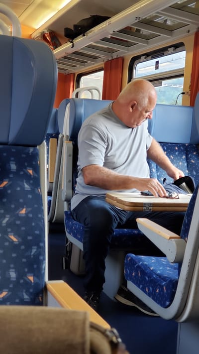 The image depicts the interior of a train carriage, likely in Margecany, Slovakia. A bald man, wearing a grey t-shirt and dark blue jeans, is seated in a blue train seat with a patterned upholstery. He is looking down and appears to be reaching into a black bag placed on a foldable wooden tray table in front of him. A pair of glasses and a mobile phone are also visible on the tray table. The train carriage features overhead storage racks with luggage, and orange curtains are drawn over the windows. The lighting suggests it is daytime.