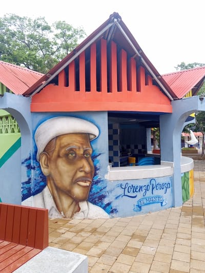 The image shows a colorful building with a mural of a man's face wearing a white cap. The mural is on a light blue wall and the man appears to be elderly with green eyes. Below the mural, the text "Lorenzo Peroza EL MAESTRO" is written in blue script. The building has an orange roof with vertical slats. The surrounding area is paved with tan square tiles, and a red bench is visible on the left side of the frame. In the background, there are trees and other structures, suggesting an outdoor public space. This mural is located on the malecón of the city of Tolú, Colombia, a popular waterfront promenade.