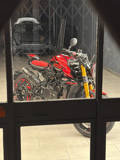 An MV Agusta motorcycle with a red and black color scheme is parked behind a glass door with a metal gate. The motorcycle is positioned at a slight angle, with its front facing left. It features prominent gold-colored front suspension forks and red accents on the wheels. An "Arrow" branded exhaust is visible. In the background, through the glass and gate, a black car with a sleek design, possibly a Ferrari, is parked. The ground is tiled with large, light-colored squares. The scene appears to be a showroom or a secure parking area. The image is taken from outside the glass door, looking in. The presence of a lock on the door suggests it is a secured entry. There are no people visible in the image. The city of Novara, Italy, is mentioned as the location, but there are no specific landmarks to confirm this.