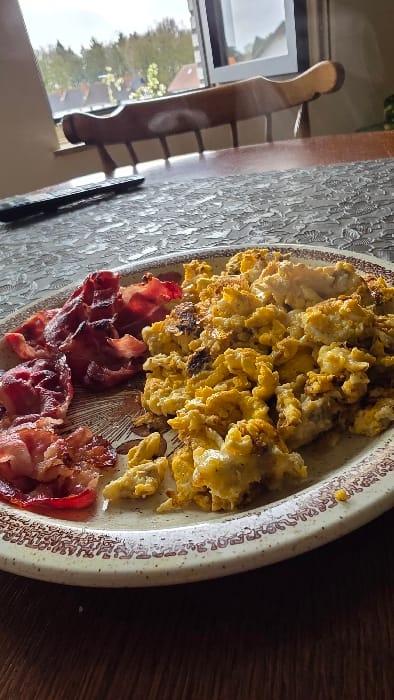 The image displays a close-up, elevated perspective of a prepared meal on a dining table within a residential setting in Heusden, Belgium. A round, off-white plate with a detailed brown border contains a serving of scrambled eggs on the right side, characterized by their yellowish hue and some browned, crispy sections. To the left, several strips of bacon are visible, varying in doneness from darker, crispier pieces to lighter, pinker sections. The plate rests on a dark brown wooden table, partially covered by a light-colored, intricately patterned tablecloth or placemat. To the immediate left of the plate, positioned on the patterned surface, lies a black remote control. In the mid-ground, behind the table, the dark brown wooden backrest of a chair with vertical slats is partially visible. Further back, a window with a white frame provides a view of an outdoor environment, showing green trees and the reddish-brown rooftops of other buildings under an overcast sky. No individuals are present in the frame.