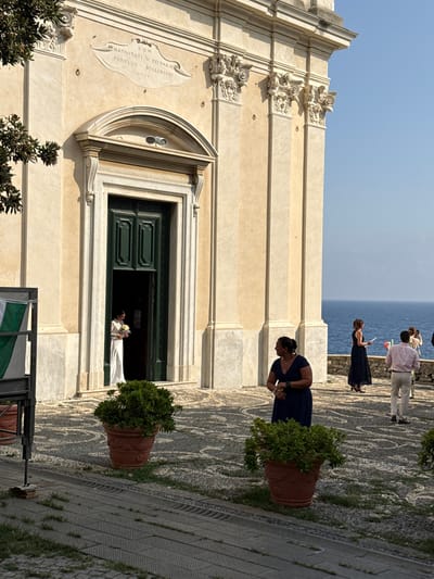 The image shows the facade of a church with a wedding party gathered outside in Bogliasco, Italy. A bride in a white dress stands in the open doorway holding a bouquet of yellow flowers. To the right of the church entrance, a woman in a dark blue dress stands looking to the left. Further to the right, near the coast, a group of people are visible, including a woman in a black dress and a man in a light pink shirt and beige pants. The church facade is decorated with classical columns and ornate carvings. The ground is paved with cobblestones, and large terracotta pots with green plants are placed in front of the church. In the background, the blue sea is visible under a clear sky. A flag with green and white stripes is partially visible on the left side of the image.