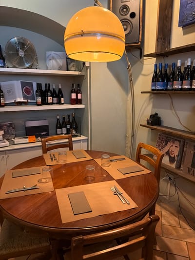 The image depicts the interior of a restaurant or wine bar, likely in Pienza, Italy, judging by the style and rustic elements. A round wooden table is set for dining with place settings that include brown paper placemats, clear glasses, and silverware. Two wooden chairs are visible at the table, one occupied by a person whose back is to the camera.

Behind the table, shelves are stocked with numerous wine bottles, some of which appear to be red and rosé. There are also books and decorative items on the shelves. A large, industrial-style fan is on a shelf to the left, and a vintage speaker system is mounted on the wall above. A hanging pendant light with a yellow shade illuminates the scene. The walls are painted a light, neutral color, and the floor is tiled. The overall impression is one of a cozy, rustic establishment.
