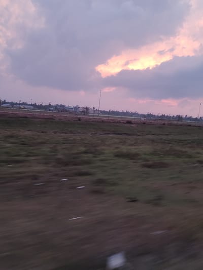 A panoramic view captures a dusky sky over an industrial area in Glokaron, Indonesia. The sky is a canvas of soft purples and pinks, with a sliver of bright orange light peeking through a band of clouds. In the mid-ground, industrial structures, possibly a port or factory, are visible against the horizon, with cranes and buildings dotted across the landscape. A faint line of palm trees separates the industrial zone from the foreground. The foreground itself is a blur of green and brown, suggesting movement, perhaps a vehicle passing by, with streaks of light across the grassy terrain. The scene is characterized by a hazy, twilight atmosphere.
