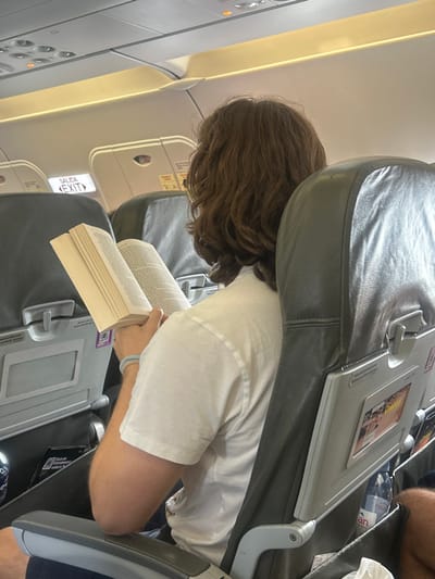 The scene is captured inside a commercial aircraft, consistent with air travel to or from San José del Cabo, Mexico.
A single individual, viewed from behind, occupies a gray upholstered passenger seat. The subject has medium-length, wavy brown hair and wears a white, short-sleeved t-shirt. A light blue and white band-style bracelet is visible on their left wrist. Both hands hold open a thick, light-colored book, indicating the subject is engaged in reading. The text within the book is not legible.
The immediate environment consists of standard airline seating. The seat directly in front of the subject features a gray plastic frame for a seat-back pocket, containing a crumpled white item, a clear plastic bottle of Evian water, and a printed publication with a partial image. A purple and white QR code sticker is affixed to the seat back. To the subject's left, another gray seat is visible, and above it, an illuminated "SALIDA / EXIT" sign is clearly displayed in both Spanish and English. Overhead, the aircraft's ceiling panel shows recessed lighting strips, air vents, and individual reading light controls. The overall cabin lighting appears bright and uniform. No other individuals are distinctly visible or interacting with the subject.