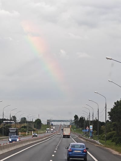 The image shows a highway with a rainbow in the sky above. Several cars and trucks are on the highway. The weather appears overcast. The scene is likely located in or near Gorodnya, Russia, given the context provided.