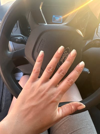 The image shows a close-up view of a person's left hand resting on a car's steering wheel. The hand has manicured nails with a light, shimmery polish. The steering wheel has a "Ford" logo in the center and "AIRBAG" text visible. The dashboard displays "31°C" and "Vehicle is on". The sun is shining brightly, casting a lens flare effect on the left side of the image, suggesting it is daytime. The person is wearing gray plaid pants. The scene is inside a car, likely in a location with warm weather. The presence of the "Ford" logo and the dashboard information provides specific cues about the car. The image is taken from a perspective that focuses on the hand and steering wheel, implying the subject is the driver. The overall mood is neutral, with no strong emotional indicators.