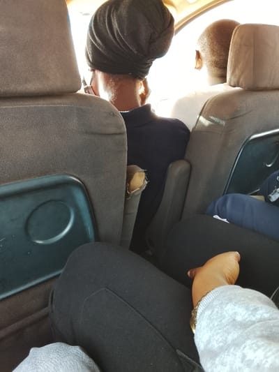 The image shows the interior of a vehicle, likely a van, with two rows of seats occupied. A person in the second row is wearing a dark head covering, possibly a turban, and a dark polo shirt. Another person in the front row is wearing a white shirt. In the foreground, a person wearing black pants and a gray sweatshirt is partially visible. An armrest between the seats is worn and torn. Seatback trays are visible on the backs of the seats. The lighting suggests it is daytime.