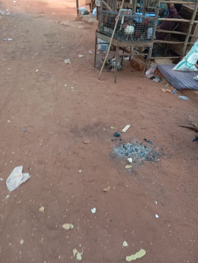 A dirt road stretches into the distance. Scraps of paper and discarded food items litter the ground. In the middle of the frame, a patch of burnt debris is visible. To the right, a wire cage houses chickens, stacked above each other. The cage is part of a stall in an outdoor market setting. A person sits behind the stall, partially obscured. Bags of goods and other market items are also present.