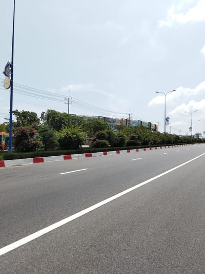 The image captures a wide, paved road with a single white lane marker extending into the distance. The road is bordered on the left by a white curb with red painted sections. Beyond the curb is a landscaped area with manicured bushes and trees, some of which are flowering in pink and purple. A tall, dark blue pole with a decorative bird emblem and a circular sign is visible on the far left. In the background, a row of multi-story buildings with colorful facades are visible. Several streetlights with dual fixtures stand along the right side of the road. The sky is overcast with some bright white clouds. A person in an orange uniform is partially visible on the far left, near a scooter. The scene appears to be in the city of Phú Thủy, Vietnam.