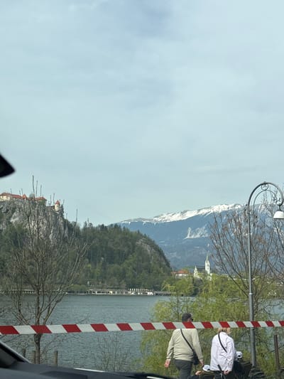 The image shows a view of Bled, Slovenia, featuring the lake and surrounding mountains. A castle sits atop a cliff overlooking the lake. A person in a beige jacket is standing near the water's edge and a person in a white jacket. A red and white barrier is visible in the foreground. In the background, snow-capped mountains rise above the landscape. A streetlight is also visible.