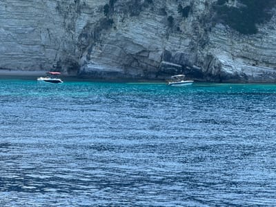 Two small boats are anchored near a rocky cliff face in the sea. The boat on the left is white with a red canopy and is closer to the shore. The boat on the right is also white and has a light-colored canopy. The sea is a vibrant turquoise color near the shore, gradually deepening to a darker blue further out. The rocky cliff rises steeply from the water, displaying layers of sediment and vegetation in crevices. The water is slightly choppy, with ripples reflecting the light. The scene appears calm and serene. There is no visible text in the image. The time of day appears to be daytime, with ample sunlight. The weather seems fair.