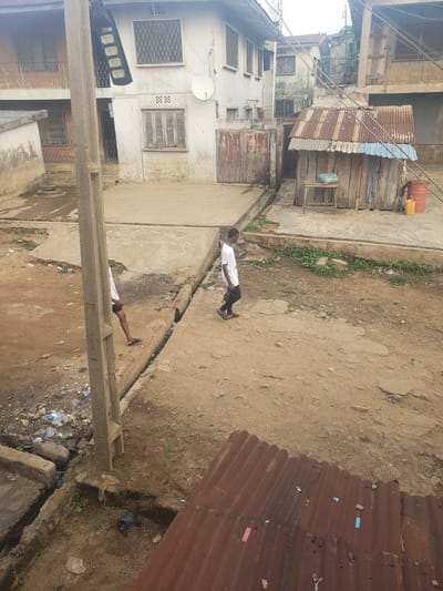 The image depicts a street scene in Alalubosa, Nigeria. The environment appears to be a somewhat impoverished urban area, evident from the weathered buildings and the presence of litter along the street. A person is walking on the street, looking at his phone, and in the foreground there is a concrete structure. Buildings are constructed of concrete and corrugated metal, some appearing rundown. The ground is primarily dirt with patches of concrete. Overhead, there are electrical wires. The overall scene suggests a developing area.